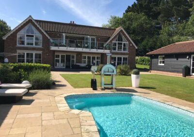 A swimming pool located in a large garden with the house to the rear. Behind the pool is a wood fired pizza oven and a stand holding utensils..